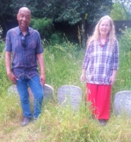 Herbie - dressed in jeans and a short sleeve shirt, with Frances, dressed in bright red trousers and checked shirt standing in the grass by a row of small gravestones